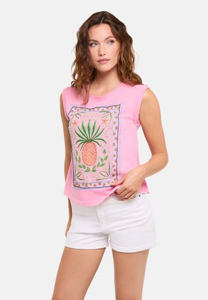 Mujer que lleva una camiseta rosa sin mangas con una gran piña bordada y pantalones cortos blancos, posando sobre un fondo liso.