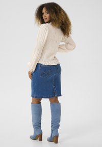 Kermakankainen smokkiblouse jossa on röyhelöhelma, yhdistettynä farkuhameeseen, ja sinisiin mokkaiseen polvimittaisiin saappaisiin joissa on kerrostettu korko.