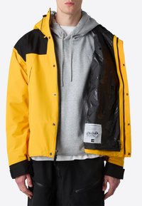 Žlutá nepromokavá bunda s černými akcenty, se zipem vpředu a patentkami. Nosená přes šedou mikinu, odhalující vzorovanou černou podšívku.
