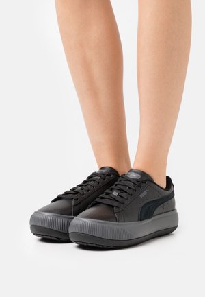 Schwarze Sneakers mit einem glatten Obermaterial aus Leder, strukturierten Akzenten und einer dicken Gummisohle. Sie verfügen über Schnürsenkel und ein Logo an der Seite.