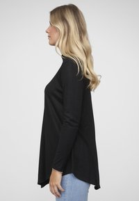 Zwarte top met lange mouwen, losse pasvorm, afgeronde zoom en zachte textuur, gecombineerd met lichtblauwe spijkerbroek. Zijaanzicht.