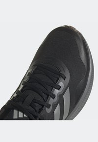 Chaussure de sport noire avec une tige en mesh, des lacets, des accents gris réfléchissants et une semelle rembourrée. Présente un design texturé et une forme profilée.