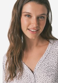 Lysegrå top med leopardmønster, med V-udskæring og dekorativ tekstaccent nær kraven. Glat stoftekstur.