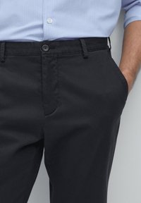 Black pants made of a smooth fabric, featuring a flat front, side pockets, and a button closure, paired with a light blue striped shirt.