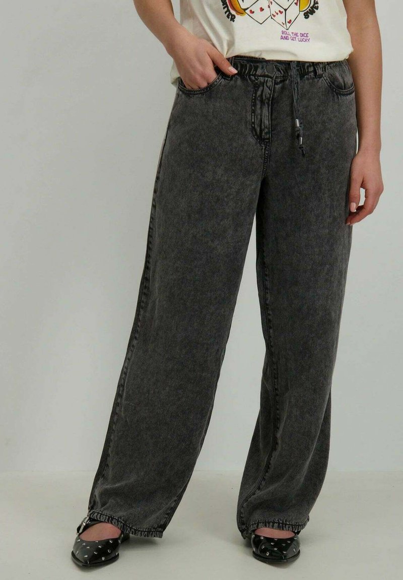 Pantalons en denim gris à jambes larges, avec une taille élastique et un cordon de serrage. Comprend deux poches et une texture légèrement délavée, associés à des ballerines noires.