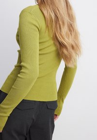 Persona con lunghi capelli biondi che indossa un maglione aderente verde a coste e pantaloni neri, in piedi contro uno sfondo chiaro e semplice.