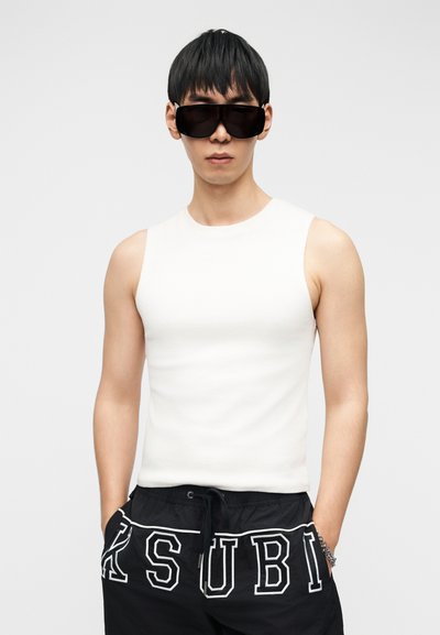 Vyras nešiojantis juodus didelius saulės akinius, baltą berankovį marškinėlį ir juodus šortus su raišteliu ir baltais užrašais, stovintis su rankomis kišenėse.