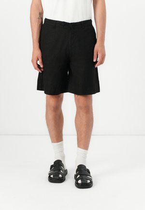 Black linen shorts with a drawstring waist, feature a loose fit and side pockets, paired with black sandals and white socks.