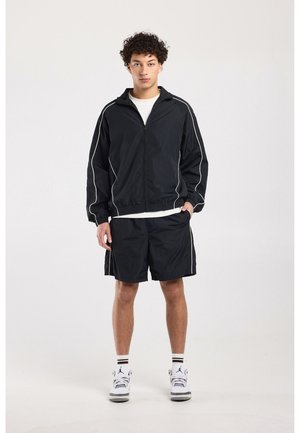 Junger Mann steht, trägt schwarze Sportjacke und Shorts mit weißen Sneakern und gestreiften Socken vor schlichtem Hintergrund.