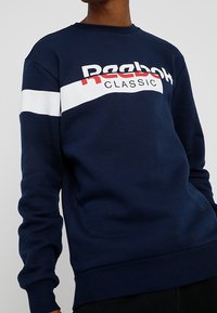 Marinblå sweatshirt med vit och röd horisontell rand med "Reebok" och "CLASSIC" branding; tillverkad av mjukt, strukturerat tyg.
