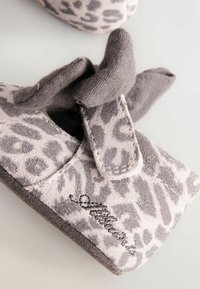 Weiche Baby-Fäustlinge aus Stoff mit grauem Leopardenmuster und einem kleinen, gestickten schwarzen Logo, liegend auf einer hellen Oberfläche.