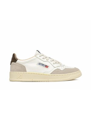Sneaker bianca basse con dettagli in pelle scamosciata beige, suola beige, lacci bianchi e una piccola toppa con logo con testo blu e una bandiera americana sul lato.