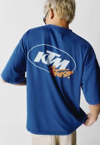 Modrá krátká tričko z bavlny s velkým bílým logem KTM a oranžovými akcenty na zadní straně. Uvolněný střih, hladká textura.