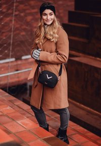 Schwarze gesteppte Umhängetasche mit rosa Logo, getragen mit einem braunen Mantel und grauen Handschuhen. Der Hintergrund zeigt Ziegelstufen und Metallgeländer.