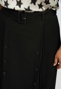 Jupe plissée noire avec une taille ajustée, dotée d'une ceinture élégante, de détails de boutons et d'un tissutexturé. Vue en gros plan de la zone de la taille.