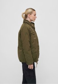 Chaqueta de estilo militar en verde oliva con un cuello alto, múltiples bolsillos frontales y cintura ajustable con cordón. Tela de algodón, ajuste relajado.