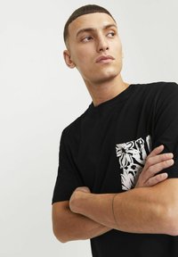 Camiseta de algodón negra con un bolsillo de patrón floral blanco en el lado izquierdo. Mangas cortas y un cuello redondo clásico.