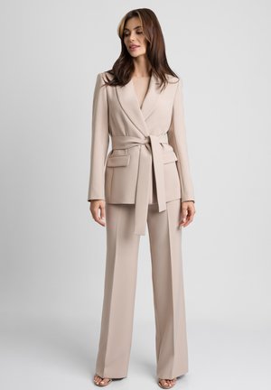 Kobieta stojąca w beżowej, paskowanej marynarce z klapowymi kieszeniami i dopasowanych szerokich spodniach na szarym, jednolitym tle.