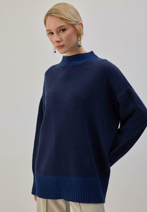 Femme blonde portant un pull ample bleu foncé avec col et ourlet côtelés, assorti à un pantalon beige clair, posant devant un fond uni.