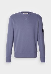 Bluza z długim rękawem i okrągłym dekoltem w stonowanym niebieskim kolorze, z prążkowanymi mankietami i dołem, z małą czarną naszywką z logo na lewym ramieniu.