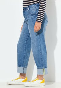 Pantalones vaqueros de mezclilla azul claro con un corte relajado, bajos doblados y ribete en azul claro contrastante. Combinados con zapatillas blancas con detalles amarillos.