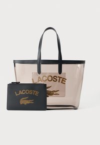 Prozirna plastična torba s crnim ručkama, koja ima središnji prozirni džep s zlatnim natpisom "LACOSTE" i logom krokodila. Uključuje odgovarajući crni džep.