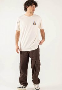 Cream short-sleeve t-shirt featuring a small graphic, paired with loose brown cargo pants and white sneakers with black accents.