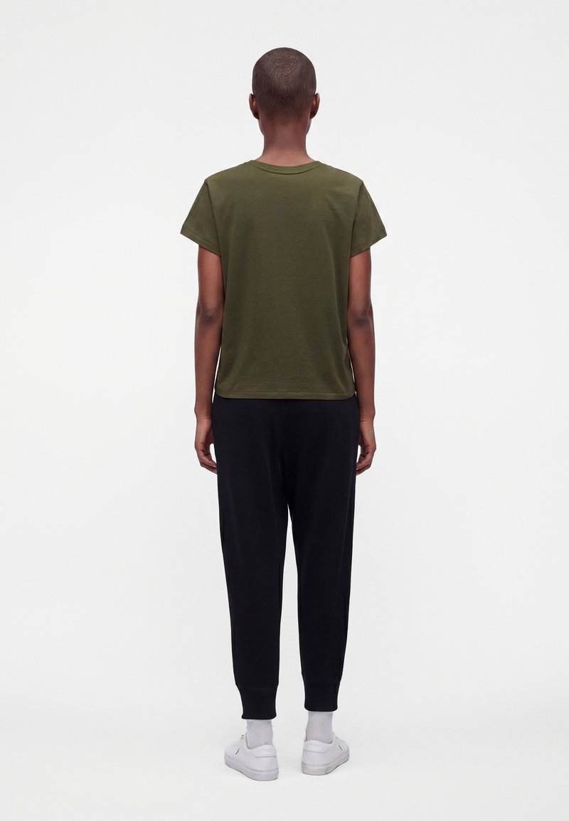 Personne aux cheveux courts portant un t-shirt vert olive, un pantalon noir, des chaussettes blanches et des baskets blanches, debout de dos.