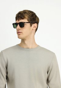 Młody mężczyzna w ciemnych okularch tortoise shell i jasnoszarej bluzie z okrągłym dekoltem, patrzący w lewo na jednolitym tle.