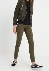 Giacca biker in suede verde oliva con cerniere argento, abbinata a una t-shirt nera e jeans skinny verde oliva. Sneakers bianco e nere completano il look.