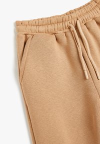 Pantalones de chándal marrón claro fabricados en una tela suave, con cinturilla elástica, cordón y bolsillos laterales; textura suave con costuras mínimas.
