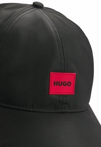 Sort kasket lavet af glat materiale med et iøjnefaldende rødt rektangulært mærke med "HUGO" i sort skrift. Designet inkluderer en rund top og en buet skygge.