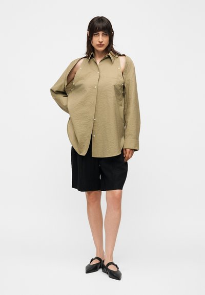 Γυναίκα στέκεται φορώντας μια φαρδιά πουκάμισο ελιάς με κουμπιά και ανοίγματα στους ώμους, μαύρα σορτς μέχρι το γόνατο και μαύρα μυτερά παπούτσια.