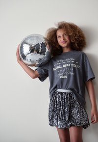 Maglietta grigia oversize con testo bianco, abbinata a una gonna plissettata nera con paillettes. In mano una palla da discoteca riflettente. Sfondo chiaro.