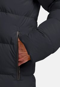 Donkerblauwe pufferjacket met een ritszak, gestructureerde stof en een gewatteerd uiterlijk. Mouw is gedeeltelijk zichtbaar.