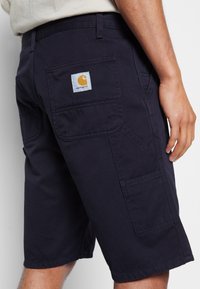 Calções azul-marinho feitos de um tecido durável, com um bolso traseiro que possui um emblema em branco e laranja da Carhartt, bolsos laterais para utilidades e pormenores cosidos.