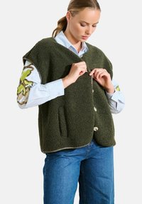 Groene fleece vest met een textuurafwerking, voorzien van een V-hals, oversized armsgaten en een voorknopsluiting. Gestyled met een blauwgestreept shirt.