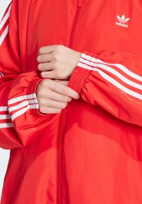 Jachetă roșie cu fermoar, cu accente albe și trei dungi albe pe mâneci. Manșete elastice și textură de material neted și lucios. Logo-ul Adidas este prezent.