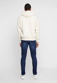 Sudadera blanca con capucha y acabado texturizado, combinada con vaqueros azul oscuro de corte ajustado y zapatillas blancas, vista desde atrás.