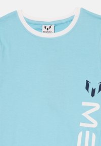 Světle modré tričko s krátkým rukávem a bílým límcem, na přední straně s námořnickým grafickým potiskem, který zobrazuje logo a stylizovaný text. Měkký bavlněný materiál.