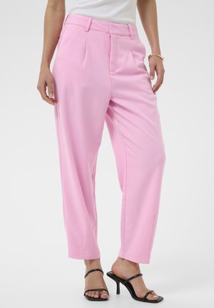 Femme portant un pantalon rose taille haute et ample, associé à des sandales à talons noires à brides et un haut blanc sans manches.