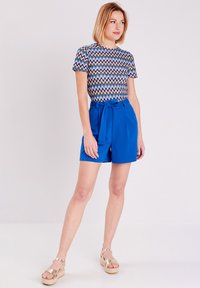 Shorts bleus avec un lien à la taille, associés à un haut à motif zigzag multicolore. Modèle se tenant sur un fond clair.