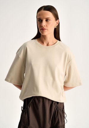 Kobieta z prostymi włosami, nosząca złote kolczyki koła, luźną beżową koszulkę i ciemnobrązową spódnicę z marszczonymi kieszeniami, stoi na białym tle.