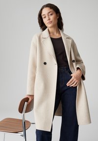 Beige wollen blendvest met een notch kraag, dubbele knopen en lange mouwen, gecombineerd met donkere jeans. Een houten en metalen stoel in de buurt.
