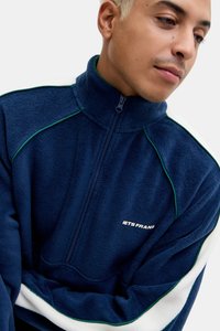 Giacca in pile blu navy con colletto alto, piping verde e strisce laterali bianche. Chiusura frontale con zip e logo sul petto. Tessuto morbido.