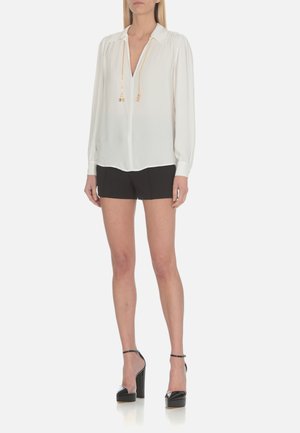 Camicia bianca a maniche lunghe con scollo a V, in tessuto leggero, con arricciature alle spalle, abbinata a pantaloni corti neri e tacchi alti.