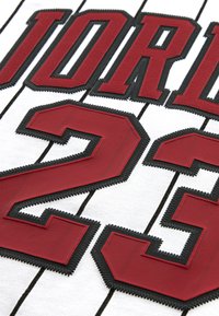 Balts sporta krekls ar sarkaniem izšūtiem burtiem "JORDAN" un skaitli "23", ar melnām šuvēm un svītrojuma rakstu.