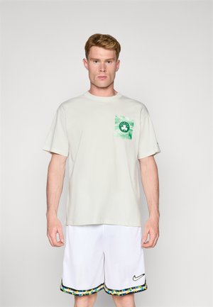 T-shirt beige clair avec une poche tie-dye verte arborant un motif de trèfle ; associé à un short blanc bordé de accents bleus et jaunes.