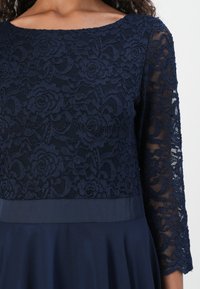Robe en dentelle marine avec des motifs floraux, dotée d'un col rond et de manches trois-quarts, complétée par une jupe en satin lisse.