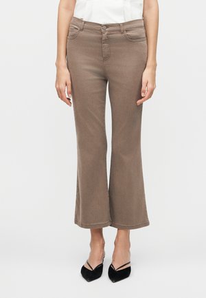 Γυναίκα που φοράει καφέ παντελόνι καμπάνα ψηλόμεσο, κομμένο στη μέση με μπροστινές τσέπες και μαύρες slingback γόβες με μυτερές μύτες, στέκεται μπροστά σε απλό φόντο.